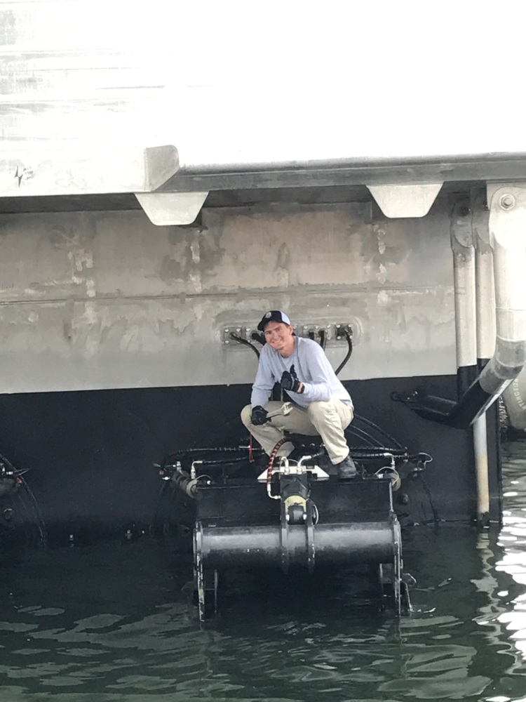 Person crouching on a platform beneath a large ship, above water.